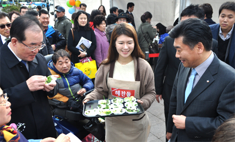 많은 사람이 모인 나트륨줄이기 국민 참여 주간 기념행사 자리에서 한 여성이 해찬들 앞치마를 두르고 시식 음식을 나눠주고 있다. 음식을 받은 사람들은 웃으며 음식을 맛보고 있다.